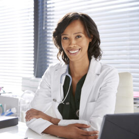 Portrait Of Smiling Female Doctor Wearing White Coat With Stethoscope Sitting Behind Desk In Office Portrait Of Smiling Female Doctor Wearing White Coat With Stethoscope Sitting Behind Desk In Office